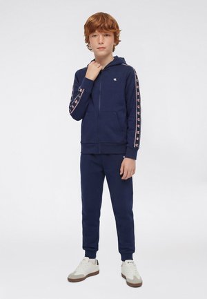 Niño pelirrojo con sudadera con capucha azul marino con cremallera y pantalones deportivos a juego con rayas de logotipo en las mangas y zapatillas blancas, de pie.