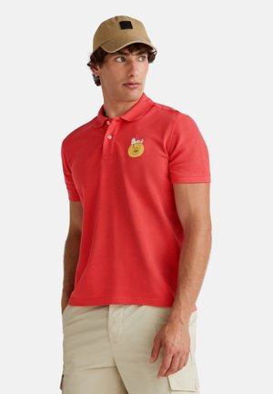 Red polo shirt with a textured finish, featuring a round yellow patch with an embroidered design. Complements with a beige cap with a black square accent.