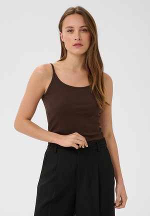 Femme aux longs cheveux bruns portant un haut marron sans manches et un pantalon noir taille haute, debout devant un fond clair uni.