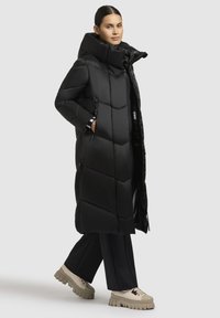 Schwarzer, langer Daunenmantel mit Kapuze, der ein gestepptes Design, seitliche Taschen und eine verstellbare Kordel hat, kombiniert mit weiteren Hosen und klobigen Schuhen.