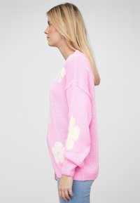 Roze gebreide trui met oversized witte bloemenpatronen, ballonnenmouwen en een ontspannen pasvorm. De textuur is zacht en lichtgewicht.