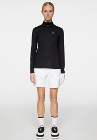 Musta pitkähiha, vetoketjullinen polo, jossa on teksturoitu kuosi, yhdistetty valkoisiin shortseihin ja golfkäsineeseen, käytetään musta-valkoisten tennareiden kanssa.