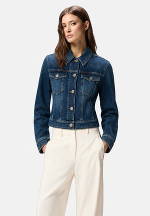 Giovane donna che indossa una giacca di jeans blu aderente con bottoni metallici e pantaloni bianchi a vita alta, mani nelle tasche, sfondo neutro.