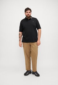 Μαύρο πόλο πουκάμισο με λογότυπο στο γιακά, σε συνδυασμό με χακί παντελόνι και μαύρα αθλητικά παπούτσια, σε στάση μπροστά σε λευκό φόντο.