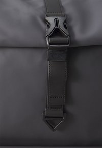 Black nylon backpack closure featuring a textured strap with a plastic buckle and rectangular metal accent labeled "TOMMY HILFIGER."