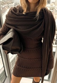 Naine, kes kannab pruuni kootud kleiti ja salli, hoides mustast nahast käekotti, seisab kõrge klaasakna kõrval, kust avaneb linnavaade.