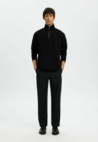 Un pull zippé noir avec un col montant, de couleur unie, associé à un pantalon noir ajusté et des chaussures noires, porté par une figure masculine debout.