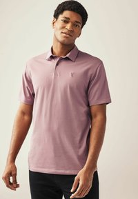 Polo de manga corta en color púrpura claro, hecho de algodón suave. Cuenta con un cuello, una tapeta de tres botones y un pequeño logo en el pecho.