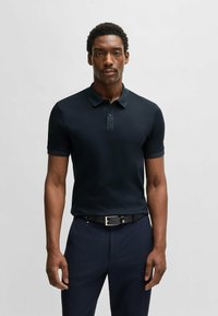 Marinblå pikétröja med korta ärmar, knäppning med tre knappar, tillverkad av ett slätt tyg. Matchad med texturerade mörka byxor och ett svart bälte.