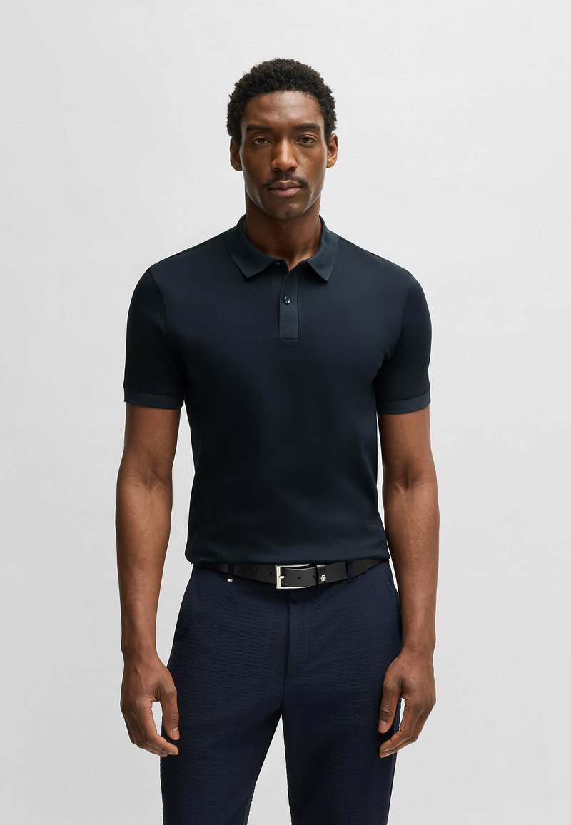 Marinblå pikétröja med korta ärmar, knäppning med tre knappar, tillverkad av ett slätt tyg. Matchad med texturerade mörka byxor och ett svart bälte.