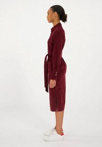 Robe en velours côtelé bordeaux avec un col, des manches longues, des fentes sur les côtés et une taille nouée. Associée à des baskets blanches avec des accents rouges.