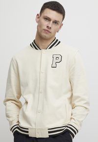 Cream varsity jacket with black striped ribbed collar and cuffs. Features "P" embroidered on the left chest. Soft, smooth fabric.