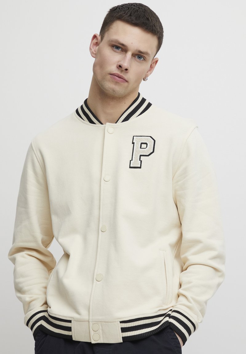 Cream varsity jacket with black striped ribbed collar and cuffs. Features "P" embroidered on the left chest. Soft, smooth fabric.