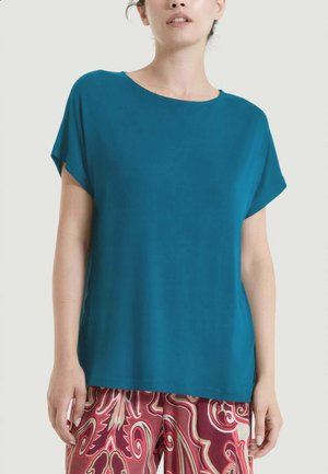 Donna che indossa una maglietta larga verde acqua a maniche corte e pantaloni rossi e beige con motivo, in piedi davanti a uno sfondo chiaro e semplice.