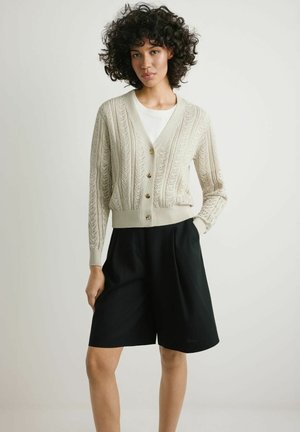 Kobieta o kręconych włosach ubrana w kremowy kardigan z warkoczowym splotem zapinany na guziki, na białym topie i czarne luźne spodenki do kolan.