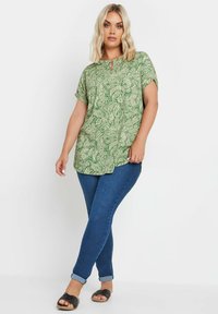 Blusa verde a fantasia con colletto a rombo, maniche corte e vestibilità comoda, abbinata a jeans skinny blu scuro e sandali neri.
