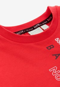 Camiseta de algodón roja con cuello redondo, texto impreso en blanco y negro, y una etiqueta de la marca en el cuello con detalles de costura blanca.