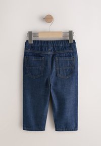 Ansicht von hinten auf dunkelblaue Baby-Jeans, die an einem Holzbügel mit Metallclips vor einem einfarbigen Hintergrund hängen.