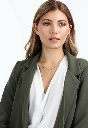 Olivgrüner Blazer über einem weißen V-Ausschnitt-Oberteil, das eine zarte goldene Halskette mit kleinen Akzenten trägt. Glatte Textur und strukturiertes Design.