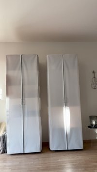 Dwie wysokie, srebrne szafki do przechowywania o gładkich powierzchniach i uchwytach. Mają minimalistyczny design i stoją przy jasnej ścianie.