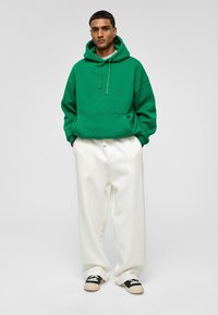 Groene hoodie met koordjes, grote voorzak, gecombineerd met losvallende witte broek en zwarte instapschoenen met een natuurlijke zool.
