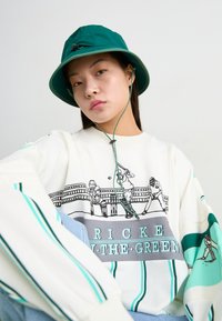 Λευκή μπλούζα φούτερ με τιρκουάζ λεπτομέρειες και γραφικό τύπωμα που απεικονίζει κριケット παίκτες. Πράσινο καπέλο με κορδόνι για το πηγούνι. Άνετη εφαρμογή.
