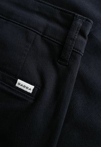 Close-up af sorte stofbukser, der viser en bæltestrop, en baglomme og et hvidt mærke med brandnavnet "GABBA".