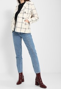 Femme portant un blazer à carreaux crème, un haut noir, un jean bleu et des bottines bordeaux brillantes, debout les mains dans les poches.