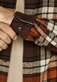 Camicia di flanella a quadri marrone e arancione con bottoni neri, dotata di colletto a piega e tessuto testurizzato, parzialmente esposta su una mano.
