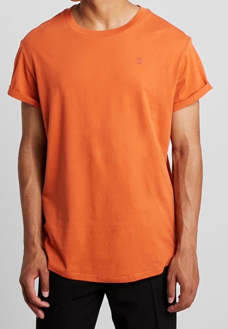 Homme portant un t-shirt orange à manches retroussées et un pantalon noir, debout devant un fond gris uni, torse visible, tête coupée.