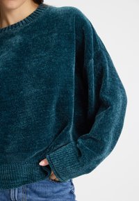 Teal színű velúr pulóver bordás nyakkal és mandzsettával, laza szabással és texturált felülettel. Farmer alsórészhez kombinálva.