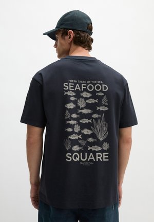 Homme portant une casquette noire et un T-shirt noir avec des illustrations de poissons et d'algues ainsi que le texte « Seafood Square » dans le dos.