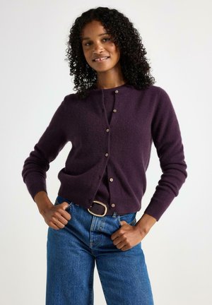 Donna che indossa un cardigan buttoned di un viola scuro e una maglietta abbinata con jeans blu, in posa con le mani nelle tasche su uno sfondo bianco semplice.