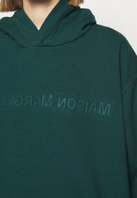 Mörkgrön huvtröja med avslappnad passform, som har texturerad "MAISON MARGIELA"-text över bröstet i matchande tyg.