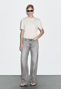 Top corto grigio chiaro a maniche corte abbinato a jeans grigi a vita alta e a gamba larga. Il modello indossa occhiali da sole scuri e semplici orecchini a cerchio.
