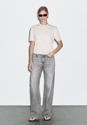 Top corto grigio chiaro a maniche corte abbinato a jeans grigi a vita alta e a gamba larga. Il modello indossa occhiali da sole scuri e semplici orecchini a cerchio.