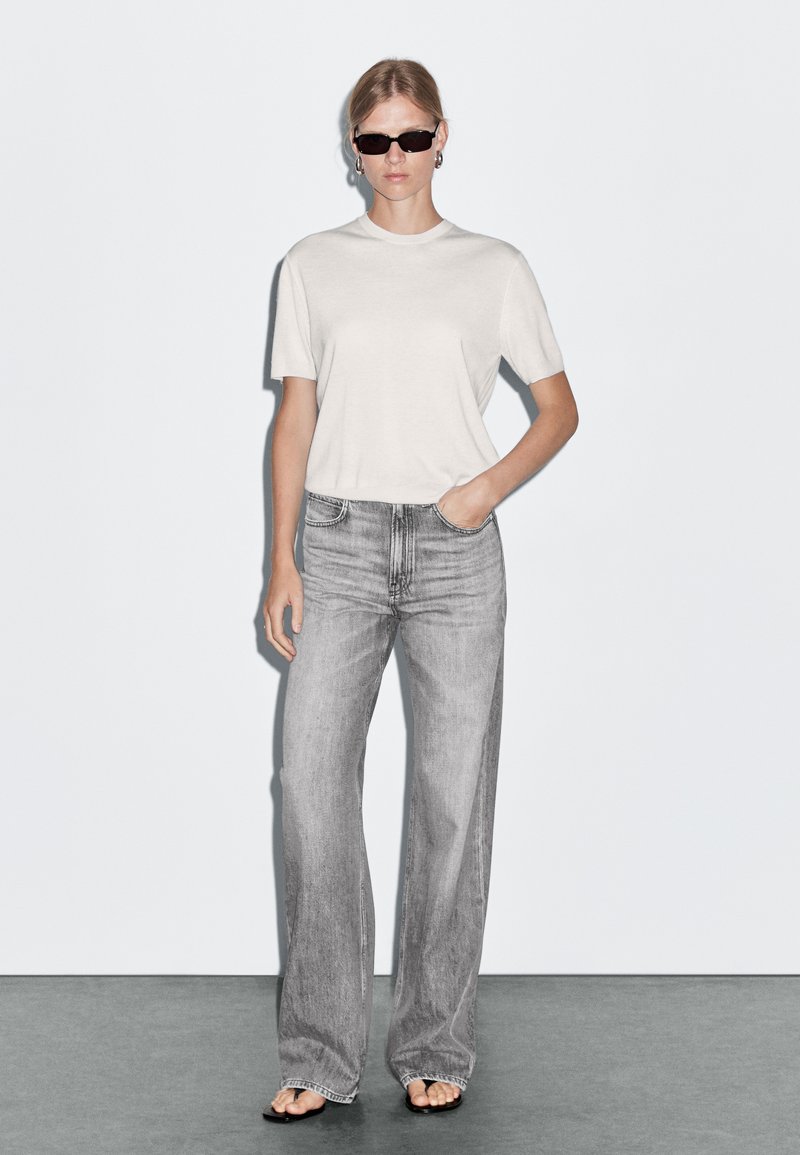 Top corto grigio chiaro a maniche corte abbinato a jeans grigi a vita alta e a gamba larga. Il modello indossa occhiali da sole scuri e semplici orecchini a cerchio.