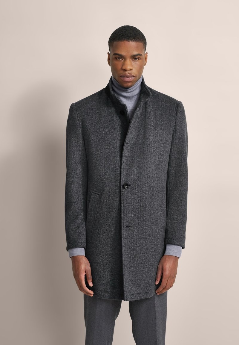 Cappotto di lana grigio con colletto, chiusura frontale a un bottone, due tasche laterali e polsini a maglia su un dolcevita grigio chiaro. Texture liscia.