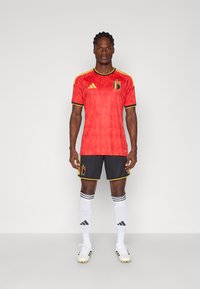 Κόκκινη πολυεστερική ποδοσφαιρική φανέλα με κοντά μανίκια, μαύρο σορτς και λευκές κάλτσες. Διαθέτει κίτρινο περίγραμμα και έμβλημα της βελγικής σημαίας.