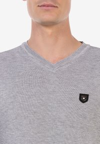 Grauer V-Ausschnitt-Pullover mit strukturiertem Strickmuster. Verfügt über ein kleines, gesticktes schwarzes Logo-Patch auf der linken Brustseite.