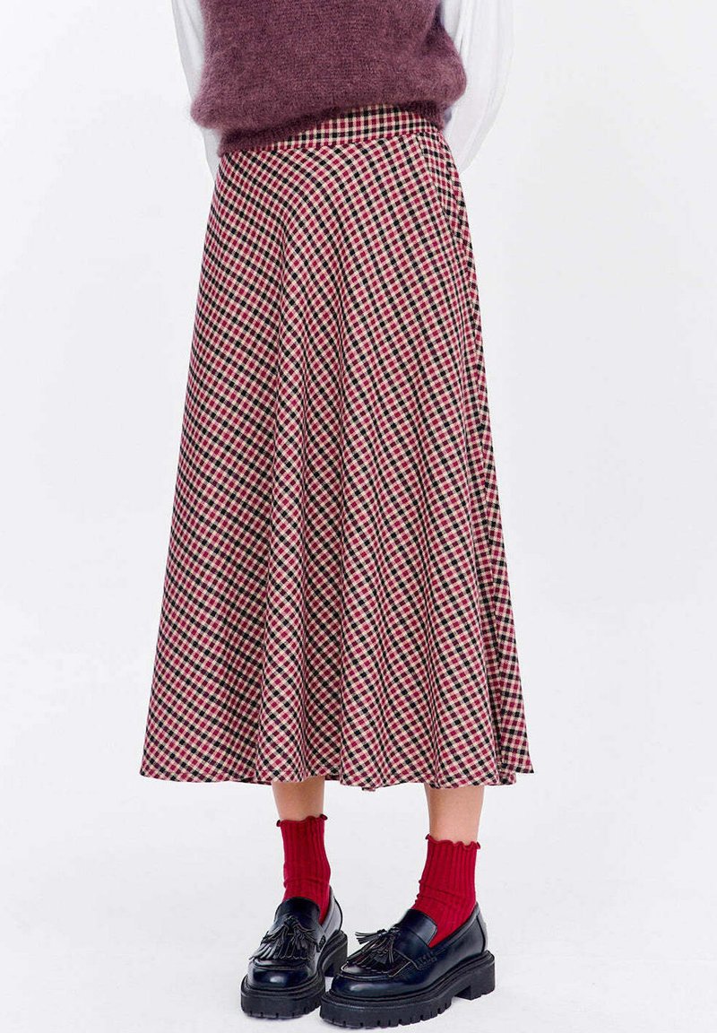 Žena oblečená v midi sukni s vysokým pásom, ktorá je károvaná v červenej, čiernej a béžovej farbe, doplnená červenými ponožkami a čiernymi mokasínami.