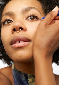 Una persona che applica l'eyeliner blu con una mano. Ha un anello al setto, pelle marrone e capelli ricci; i tratti del viso e la texture delle labbra sono visibili.