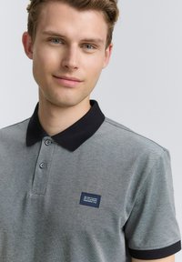 Grå polo-skjorte med sort krave, to-knaps placket og et mærke på venstre bryst. Stoffet har en tekstureret overflade.