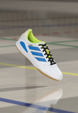 Zapatilla deportiva blanca con rayas azules, cordones negros y un forro interior verde neón. Cuenta con una parte superior texturizada y una suela de goma color chicle.