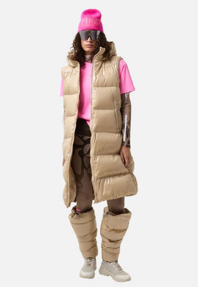 Beige polstret vest med høj krave, parret med en lys pink t-shirt, mønstrede leggings og beige puffede benvarmere. Store solbriller.