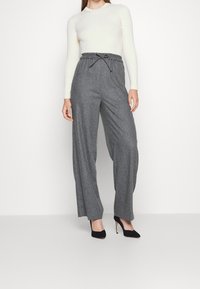 Femme portant un pantalon gris taille haute à jambes larges avec une taille à cordon, un haut blanc en maille côtelée à manches longues, et des talons hauts noirs.