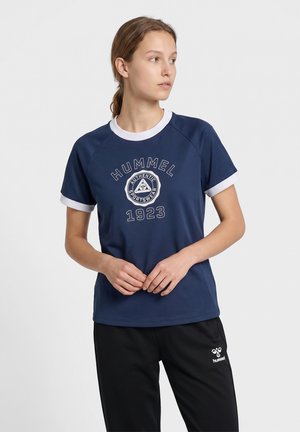 Młoda kobieta ubrana w granatową sportową koszulkę Hummel i czarne spodnie, stoi ze splecionymi rękami, patrząc w lewo.