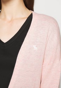 Světle růžový kardigan s měkkou texturou, s výstřihem do V a černou spodní vrstvou pod ním. Malý detail loga na kardiganu.