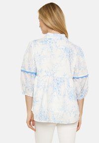 Blusa bianca con motivo floreale blu, maniche a palloncino corte e orlo arricciato. Presenta un colletto sul retro e dettagli di rifinitura blu.