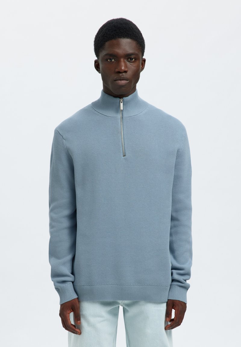 Mann trägt hellblauen Strickpullover mit halbem Reißverschluss am Kragen und hellfarbige Hose, steht vor hellem einfarbigem Hintergrund.